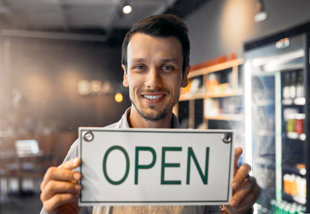 Estamos abiertos a los negocios. Retrato recortado de un joven guapo sosteniendo un cartel abierto mientras estaba de pie en su cafetería. - Foto, imagen