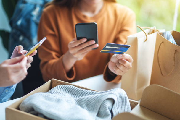 Дві жінки використовують мобільний телефон та кредитну картку для онлайн-покупки з сумкою для покупок та поштовою посилкою одягу на столі
 - Фото, зображення