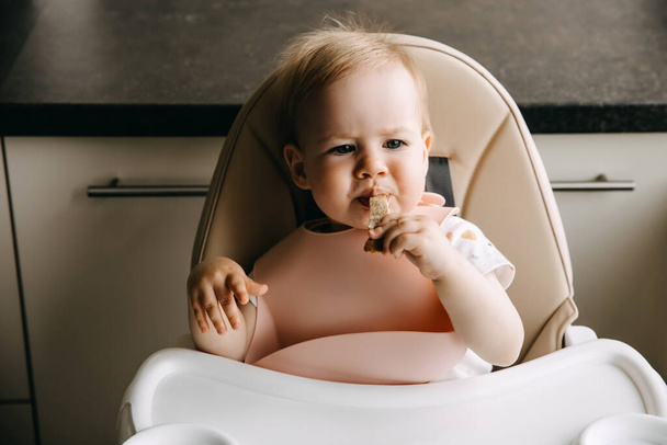 Baby sitting in high chair, with a silicone bib, eating bread. - 写真・画像
