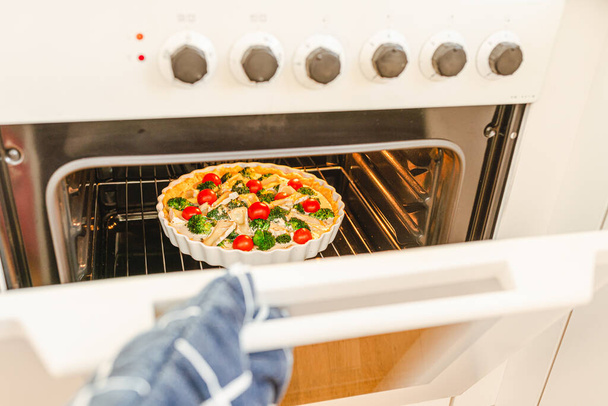 Gehen Sie in den Ofen und probieren Sie hausgemachte Quiche Lorraine mit Huhn, Brokkoli und Tomaten. Hand in Hand öffnet Backofen, wo ein offener Kuchen für den Urlaub gebacken wird. - Foto, Bild