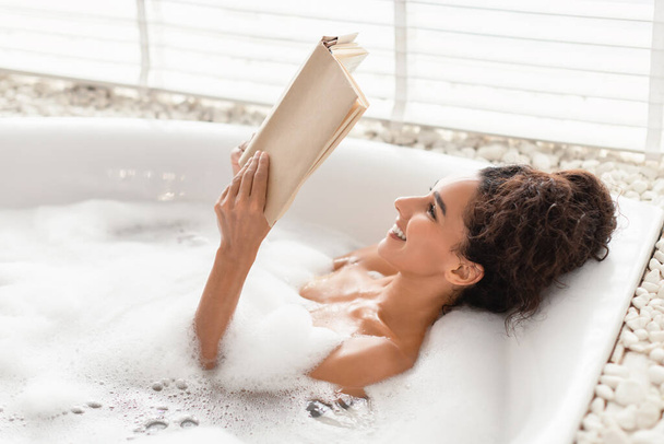 Urocza młoda kobieta leżąca w wannie z bąbelkami, czytająca książkę w relaksującej atmosferze w domu, wolna przestrzeń - Zdjęcie, obraz