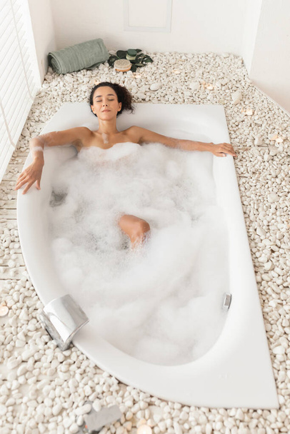 Mooie jonge vrouw liggend in bad met gesloten ogen, genieten van een ontspannen bad in luxe spa, bovenaanzicht - Foto, afbeelding
