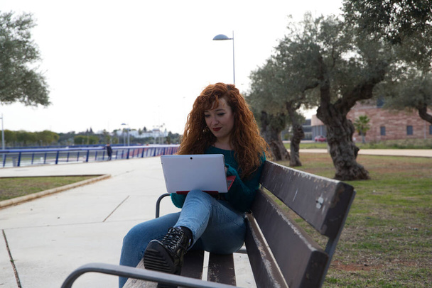 jonge en mooie roodharige vrouw zit op een bankje in een park waar veel olijfbomen staan. Ze kijkt op haar laptop naar sociale netwerken en werkt als een invloed. Begrip studenten. - Foto, afbeelding