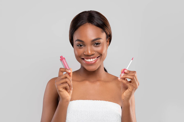 Ευτυχισμένη όμορφη νεαρή αφροαμερικάνικη γυναίκα με τέλειο δέρμα σε πετσέτα κρατήστε το lip gloss και πινέλο - Φωτογραφία, εικόνα