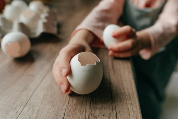 Little girl holding an egg and an empty eggshell in her hands showing how to peel an egg. Preparation for planting seeds. Creative DIY idea for gardening with kids at home. - Foto, Imagen