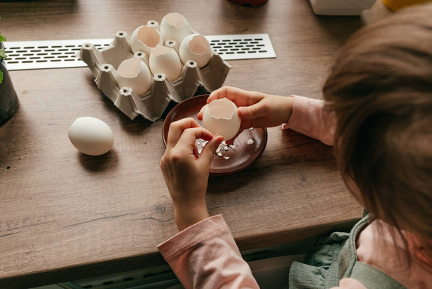 Sluiten van een klein meisje schil een ei voor het planten van zaden in een eierschaal. Huis tuinieren met kinderen op de keuken vensterbank. Selectieve focus. - Foto, afbeelding