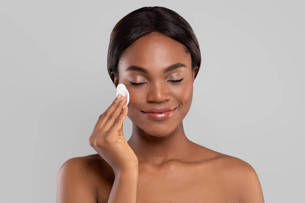Sorrindo muito jovem senhora afro-americana com pele perfeita e olhos fechados limpando seu rosto com algodão pad, - Foto, Imagem
