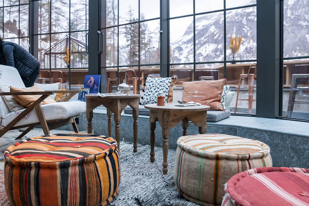 Colorful ottomans by stools and chair. Furniture arranged in stylish dining area. View of majestic mountain from glass window at luxurious holiday home. - Photo, Image