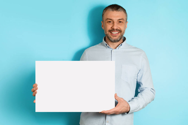 Χαρούμενος μεσήλικας άνδρας που κρατά κενό χαρτί αφίσας, μπλε φόντο - Φωτογραφία, εικόνα