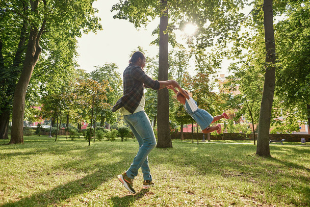 Black father spinning his pleased daughter on lawn - Φωτογραφία, εικόνα