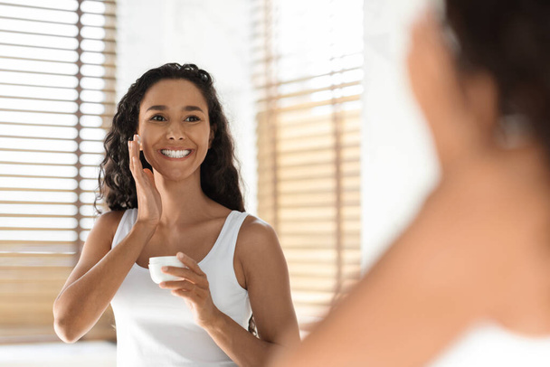 Concepto de cuidado de la piel. Bastante sonriente mujer aplicando crema facial hidratante en el baño - Foto, Imagen