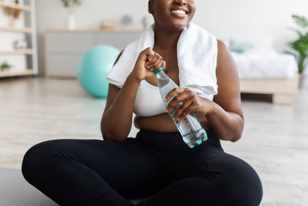 Onherkenbare overgewicht jonge zwarte dame zitten op sportmat met fles helder water thuis, close-up - Foto, afbeelding