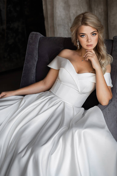 Happy feminine and stylish blonde bride posing near a large window, good mood. The girl in the chair - Foto, imagen