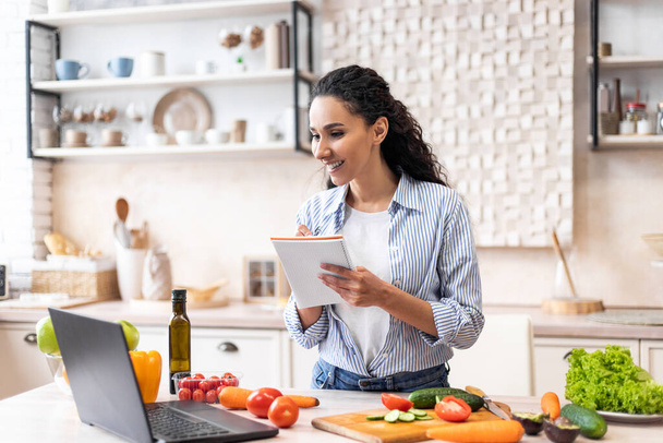 Mulher positiva escrevendo nova receita enquanto assiste vídeo no laptop, cozinhar no interior da cozinha moderna - Foto, Imagem