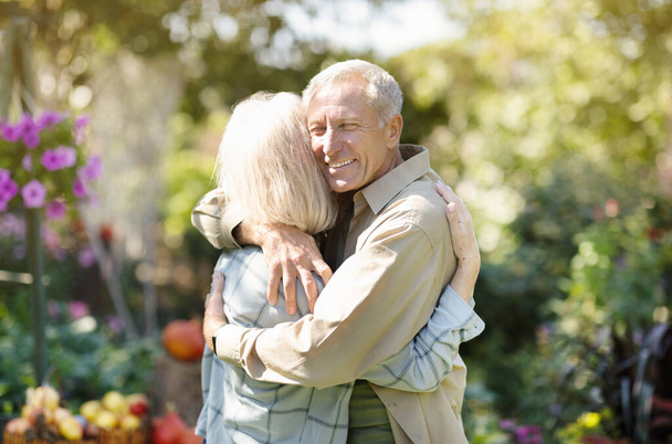 Loving elderly couple hugging each other during walk in their own garden in countyside, enjoying their vacation - 写真・画像