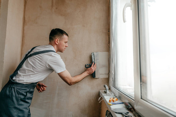 Young man builder dressed in work clothes overall applying putty mortar on the balcony wall with a spatula. - Photo, Image
