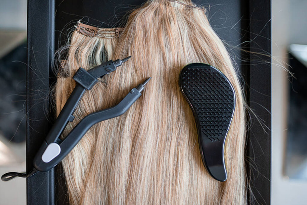 Μια σειρά από διάφορα εργαλεία επέκτασης μαλλιών για μια ξανθιά γυναίκα σε ένα σαλόνι ομορφιάς. Τσιμπίδα και μικροσωλήνες. - Φωτογραφία, εικόνα