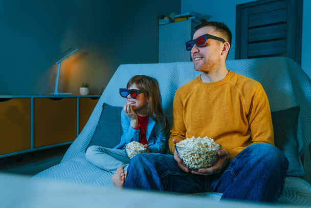 Father and daughter watching movie at home using 3D eye glasses and eating popcorn. Family activities. Selective focus on the male face. Blue light from the screen. - Foto, Imagen