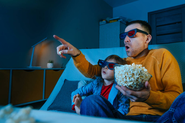 Father and daughter watching movie at home using stereo 3D eye glasses and eating popcorn. Family activities. Selective focus on the male face. Blue light from the screen. - Photo, image