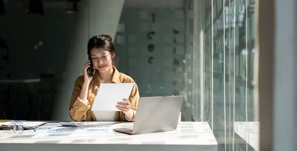 Young asian accountant calling marketing consultants and using laptop computer to analyze sales growth in the global workplace market. accounting concept - 写真・画像