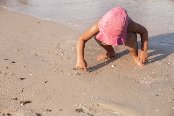 Menina criança em um chapéu de panamá rosa desenha na areia na praia - Foto, Imagem