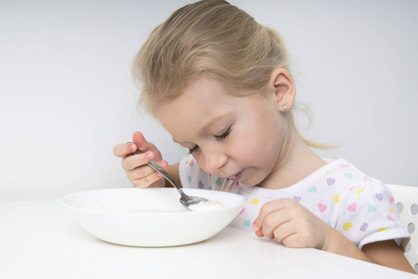 triest kind meisje blond eet pap aan een witte tafel - Foto, afbeelding