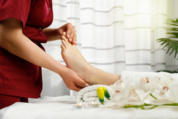 Cropped side view photo of a master in red uniform making feet massage to a woman client in spa salon, relax procedure concept. - Photo, image