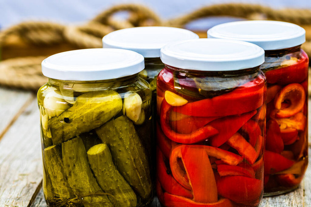 Glass jars with pickled red bell peppers and pickled cucumbers (pickles) isolated. Jars with variety of pickled vegetables. Preserved food concept in a rustic composition. - Photo, Image