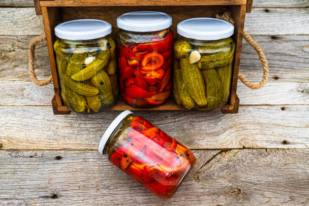 Glass jars with pickled red bell peppers and pickled cucumbers (pickles) isolated in wooden crate. Jars with variety of pickled vegetables. Preserved food concept in a rustic composition. - Photo, Image