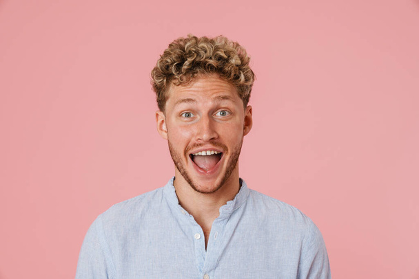 Young blonde man with curly hair expressing surprise at camera isolated over pink wall - Fotoğraf, Görsel