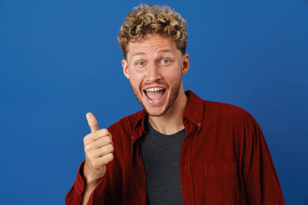Young blonde man with curly hair screaming and showing thumb up isolated over blue wall - Fotoğraf, Görsel