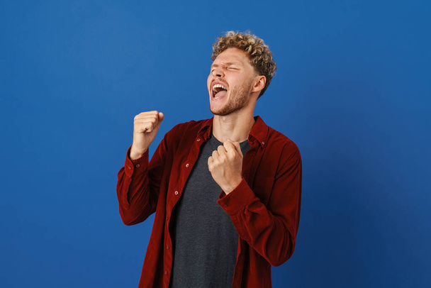 Young blonde man screaming while making winner gesture isolated over blue wall - Fotografie, Obrázek