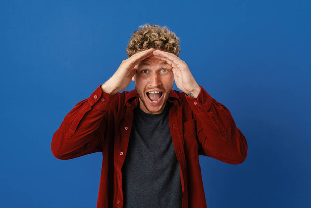 Young blonde man with curly hair expressing surprise at camera isolated over blue wall - Fotografie, Obrázek
