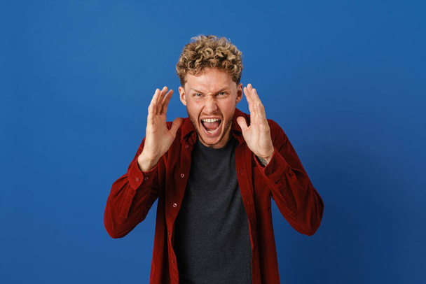 Young blonde man grimacing while screaming at camera isolated over blue wall - Foto, Bild