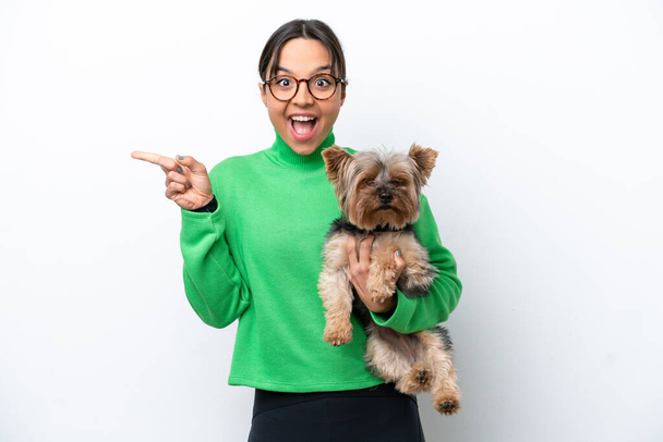 Young hispanic woman holding a dog isolated on white background surprised and pointing finger to the side - Foto, imagen