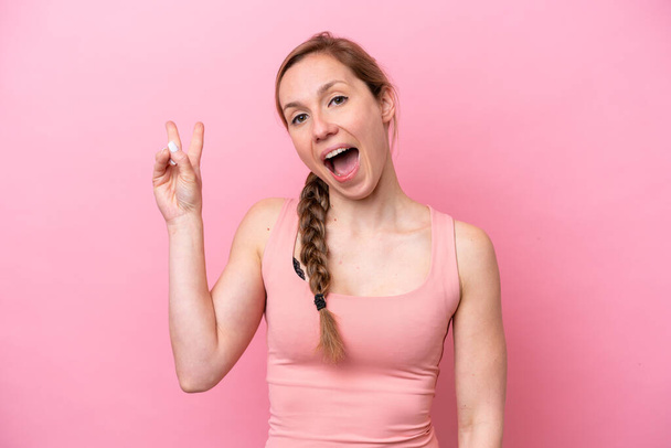 Young caucasian woman isolated on pink background smiling and showing victory sign - Foto, afbeelding