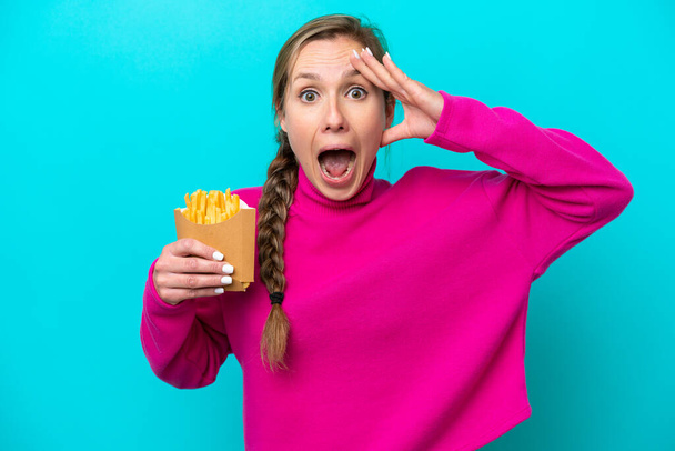 Молода біла жінка тримає смажені чіпси ізольовані на синьому фоні з несподіваним виразом
 - Фото, зображення