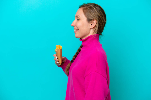 Молода біла жінка тримає смажені чіпси ізольовані на синьому фоні, сміючись у бічному положенні
 - Фото, зображення