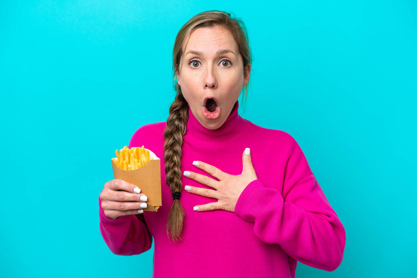 Молода біла жінка тримає смажені чіпси ізольовані на синьому фоні здивовані та шоковані, дивлячись праворуч
 - Фото, зображення