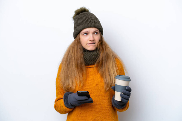 Teenager Russian girl wearing winter jacket isolated on white background holding coffee to take away and a mobile while thinking something - 写真・画像