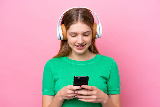 Teenager Russian girl isolated on pink background listening music and looking to mobile - Zdjęcie, obraz