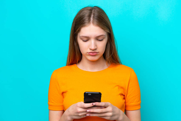 Teenager Russian girl isolated on blue background using mobile phone - Φωτογραφία, εικόνα