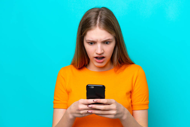 Nastolatka Rosjanka odizolowana na niebieskim tle patrząc w kamerę podczas korzystania z telefonu komórkowego z zaskakującym wyrazem - Zdjęcie, obraz
