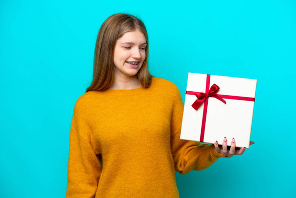 Teenager Russian girl holding a gift isolated on blue background with happy expression - Fotografie, Obrázek