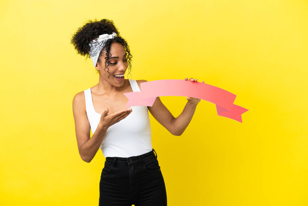 Young african american woman isolated on yellow background holding an empty placard with surprised expression - Foto, Imagen