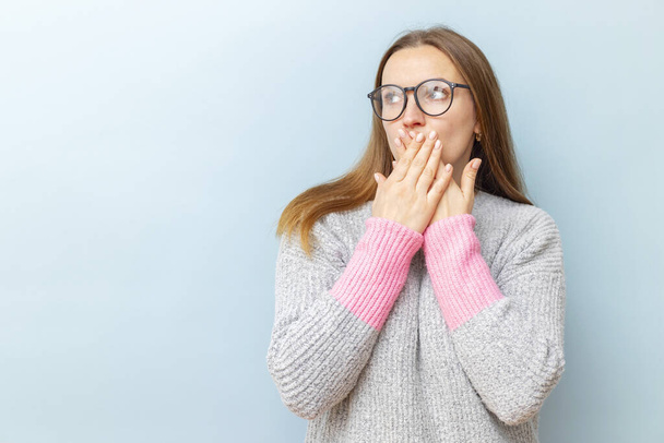 Shocked young woman in eyeglasses covering her mouth with her hands and looking at empty space for your text or object on blue background. - Фото, изображение