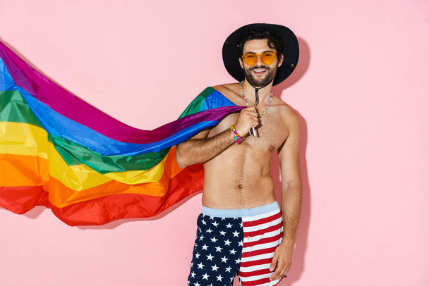 Shirtless man in hat laughing while posing with rainbow flag isolated over pink wall - Photo, image