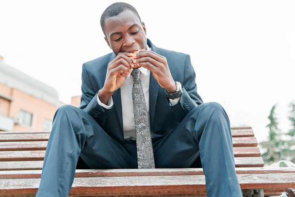 african-american man eating burger in a park outdoors while using his smartphone - Foto, imagen