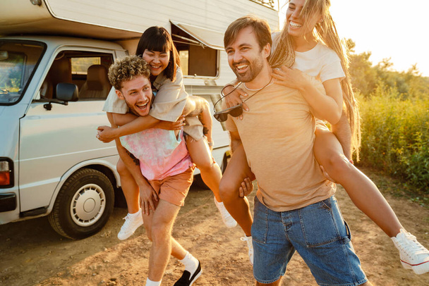 Multiracial two couples laughing and piggybacking by trailer outdoors - Foto, immagini