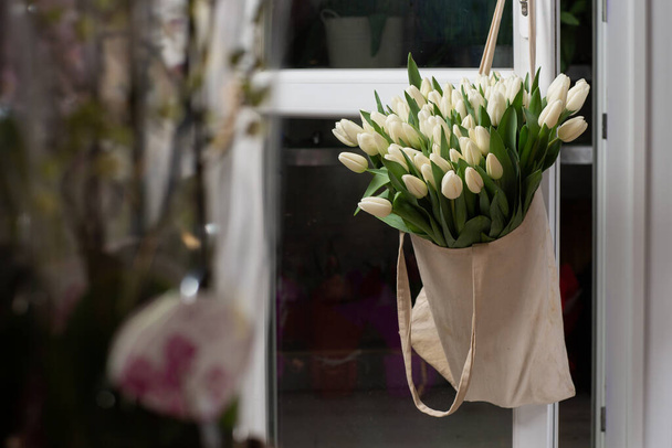 Composition with beautiful fresh flowers white tulips in eco bag hanging on door handle. Template for print. Spring shopping. - 写真・画像